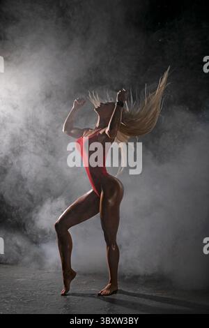 blonde female athlete displays her strength and confidence while performing in smoky gym environment, emphasizing athleticism and determination Stock Photo