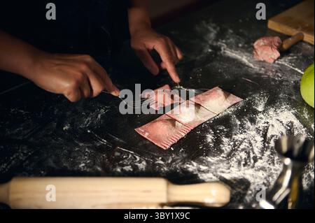 Homemade cooking scene, a woman designs ravioli using a fork on red pasta dough, evoking Italian culinary traditions and flavors. Floured tabletop and Stock Photo