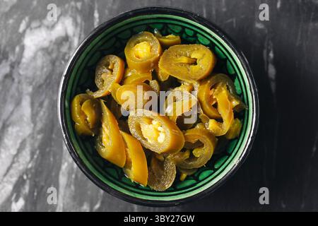 Pickled jalapeno chili slices in ceramic bowl. Spicy green peppers on dark marble background. Tangy preserved vegetable condiment for cooking or snack Stock Photo