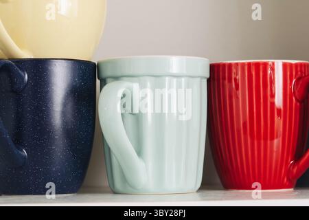 Colorful ceramic mugs on kitchen shelf. Close-up of four coffee cups in pastel and vivid colors. Modern pottery collection in cozy home interior. Stock Photo