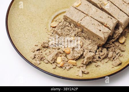 Sliced halva with peanuts on ceramic plate. Sweet crumbly dessert made of sesame paste. Middle Eastern traditional treat. Isolated on white background Stock Photo