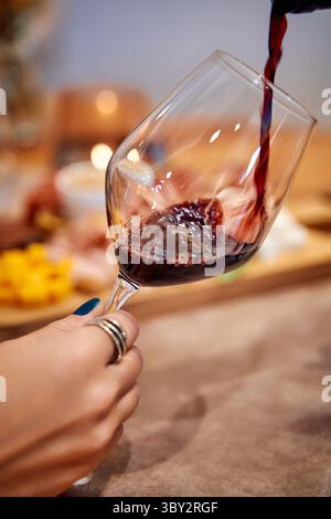 A hand elegantly pours red wine into a tilted glass with a charcuterie board in the background, showcasing a dining and entertaining setting. Stock Photo