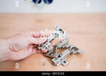 Close-up of carpenter's hand holding metal hinges, preparing to install them on wooden furniture, demonstrating precision and craftsmanship in a workshop environment Stock Photo