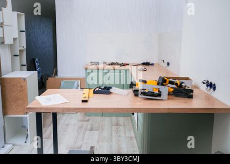 Carpenters are assembling a new kitchen using tools like level, electric screwdriver and jigsaw placed on a wooden countertop, with furniture parts and blueprint visible Stock Photo