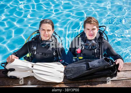 Two scuba divers standing in a pool, looking at the camera, smiling with their fins in front of them on the deck. Wearing full wetsuits and scuba gear Stock Photo
