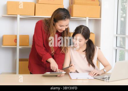 Female colleagues or partners efficiently manage and discuss online orders using a tablet and laptop in their home-based e-commerce workspace. Stock Photo