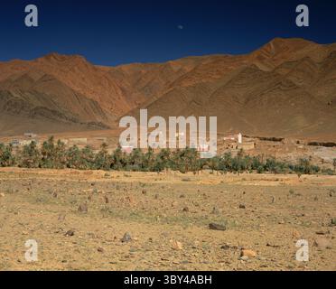 The moon rises over mountains behind a village and palm grove in remote Central Morocco with stones marking a Berber cemetery in the foreground. Stock Photo