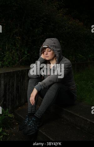 Woman with hood sits depressed on stairs, rests head, looks down Stock Photo