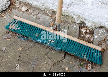 Close-up of a teal push broom with a wooden handle resting on a cracked concrete patio next to a rough concrete wall. Stock Photo