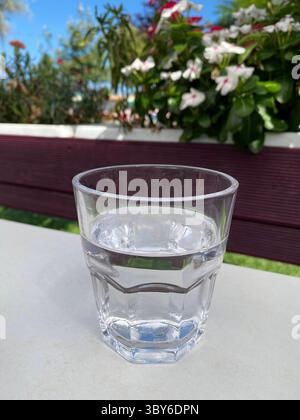 Clear, faceted glass filled with water, placed on a light-colored, outdoor table. The water inside appears still and clear, reflecting some light. Stock Photo