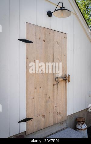 Close-up of a weathered wooden barn door with black hardware and a farmhouse-style gooseneck light fixture mounted above. A single rusted milk can res Stock Photo