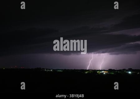 Dramatic night scene with multiple lightning bolts striking from dark storm clouds over a distant city skyline. Long exposure shot with glowing city l Stock Photo