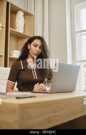 A young woman with curly hair works at the desk using a laptop. She was distracted from the screen and looks thoughtfully at the camera Stock Photo