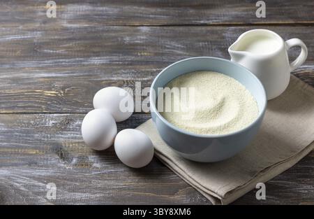 Food, Cooking ingredients featuring a blue ceramic bowl filled with semolina, three fresh white eggs, and a white jug of cream on rustic wooden table Stock Photo