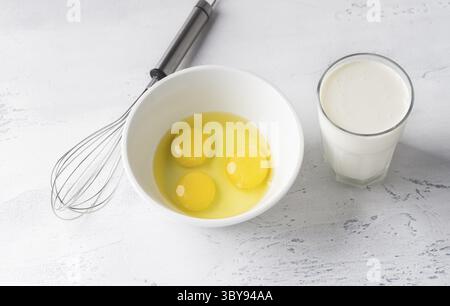 Food, White bowl with three broken eggs without shell, a glass of cream or milk and a whisk for whisking on a light gray background, top view. Cooking Stock Photo