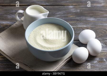 Food, Cooking ingredients featuring a blue ceramic bowl filled with semolina, three fresh white eggs, and a white jug of cream on rustic wooden table Stock Photo