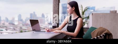 Empowered Asian adult woman confidently works at urban rooftop location She plans career goals enjoying modern city views Stock Photo