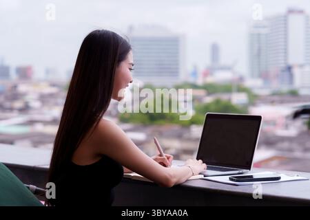 Adult Asian woman confidently works using technology. Focused on her laptop writing essential notes from an urban outdoor location. Stock Photo