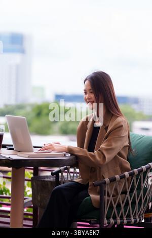 Smiling Asian woman engages in flexible work on her computer from a beautiful outdoor cafe. She embodies contemporary professional success and indepen Stock Photo