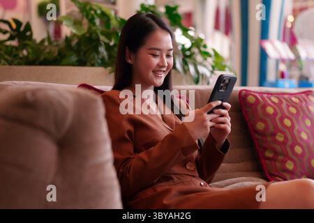Modern Asian businesswoman happily engages with her smartphone in a stylish lounge area. She exudes confidence connecting digitally with ease. Stock Photo