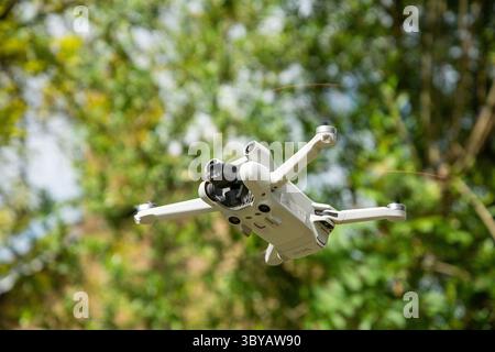 Mini drone captured in mid-flight with a backdrop of green trees, showcasing modern aerial technology in a natural outdoor environment. Stock Photo