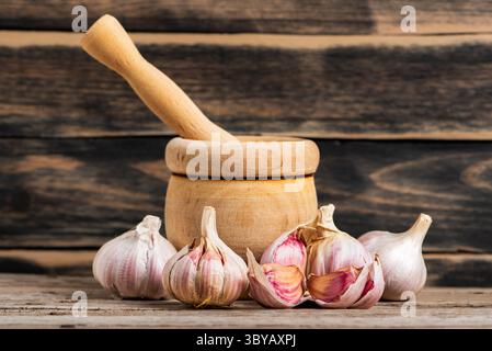 Garlic bulbs and cloves next to a wooden mortar in the foreground, with an old dark wooden background. Front view. Stock Photo