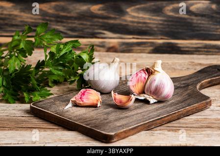 Garlic bulbs and cloves on a cutting board next to parsley sprigs, on a wooden background. Close-up. Stock Photo