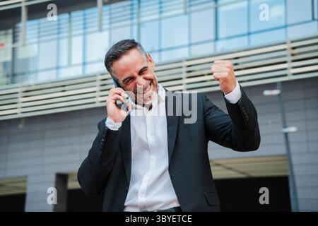 A joyous businessman expressing excitement after a successful phone call, showcasing his professional victory and determination in a well-tailored Stock Photo