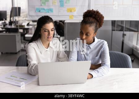Two women colleagues sit at desk in office using laptop Stock Photo