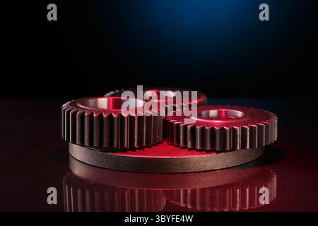 Three metallic gears rest on a dark red surface, illuminated by a blue light. The gears are a deep red color with a brushed finish. Reflections of the Stock Photo