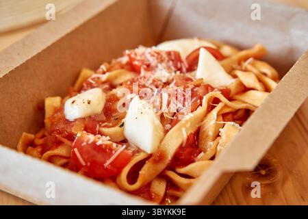 Fresh tagliatelle pasta served in a cardboard box, topped with mozzarella cheese, juicy tomatoes, and grated herbs for a delicious italian dish. Stock Photo