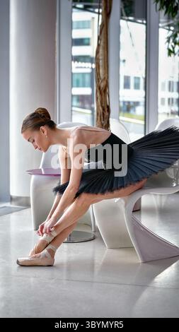 Ballerina adjusting pointe shoes while sitting on modern chair Stock Photo