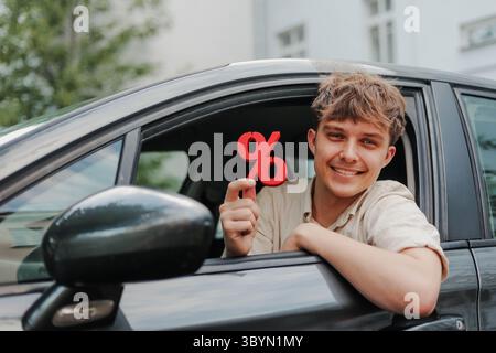 Happy young man sitting inside his car holding red percentage symbol Stock Photo