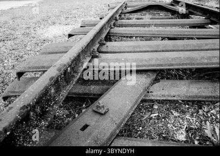 Verrostete Bahngleise im Detail Detailaufnahme eines stillgelegten Weichenstücks in Schwarzweiß. Die massiven, rostigen Stahlprofile liegen ungenutzt auf dem Boden, Spuren von Verfall und Zeit sind deutlich sichtbar. Ein Symbol für den Rückbau alter Bahnstrecken und den Wandel der Verkehrsinfrastruktur. *** Rusted railroad tracks in detail Close-up of a disused section of switch in black and white The massive, rusty steel profiles lie unused on the ground, traces of decay and time are clearly visible A symbol of the dismantling of old railroad lines and the transformation of transport infrastr Stock Photo