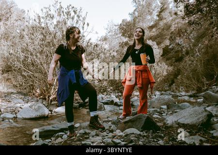 Two female hikers are crossing a river while laughing and enjoying their hike in the forest Stock Photo