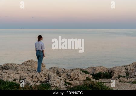 Young woman stands on a rocky coast, gazing out at the tranquil ocean during a serene sunset Stock Photo