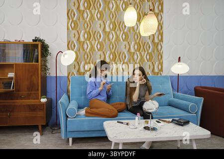 Two young women are engaged in a casual conversation about makeup while sitting on a stylish blue couch in a living room Stock Photo