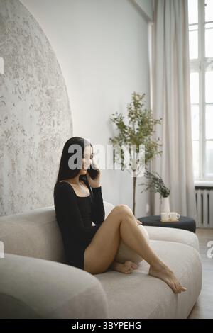 A young woman dressed in a black outfit sits comfortably on a light-colored couch Stock Photo