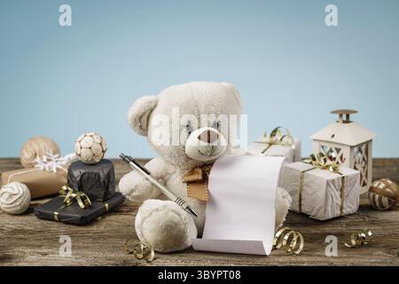 White or light brown adorable teddy bear with empty shopping or wish list and shiny, glittering christmas decorations and gifts. Holiday concept with Stock Photo