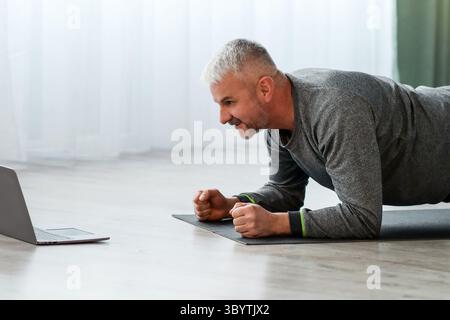 Athletic bearded older man doing exercises at home, using laptop Stock Photo