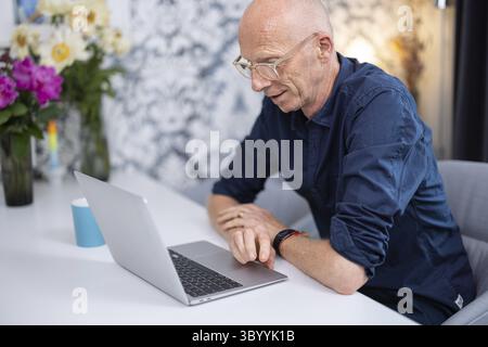 Elderly, bald man sitting in his living room at a table with flowers and a cup of coffee, working on his laptop, Cologne, North Rhine-Westphalia, Germ Stock Photo