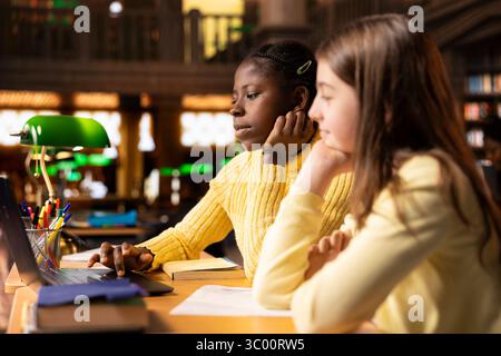 Two schoolgirls in the library participate at an online class webinar, using technology to connect with their teacher for an interactive virtual lesson. Remote courses via video call. Stock Photo