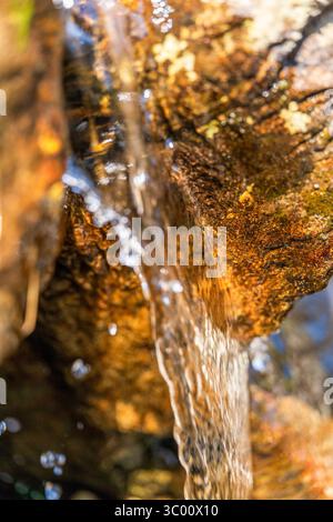 Veiled cascade of clear water slipping over rust-toned bedrock, each droplet and ripple catching golden light in intimate relief Stock Photo