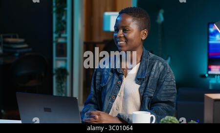 Portrait of jolly woman sitting on chair at home office desk in neon illuminated apartment late at night. Cheerful person in RGB lit living room enjoying leisure time, camera A Stock Photo