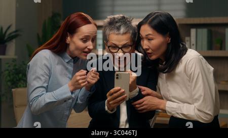 Happy excited women colleagues team group holding mobile phone multinational read good news businesswomen winning online finance celebrating office Stock Photo