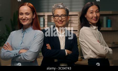 Three confident multiethnic women business office Caucasian Asian colleagues looking at camera females smiling multiracial posing crossed arms hands Stock Photo