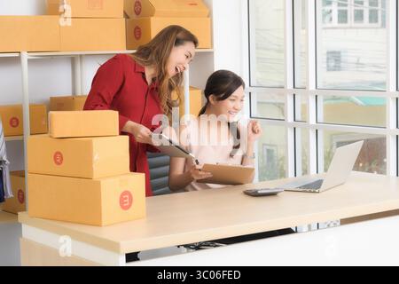 Two smiling Asian women, business partners, happily discuss operations and review documents for their online store in a bright home office setting. Stock Photo