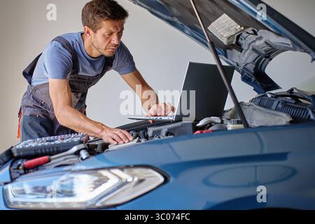 Auto mechanic using laptop for diagnostics vehicle engine in auto repair shop. Man in work overalls standing near car with open hood in garage. Vehicl Stock Photo