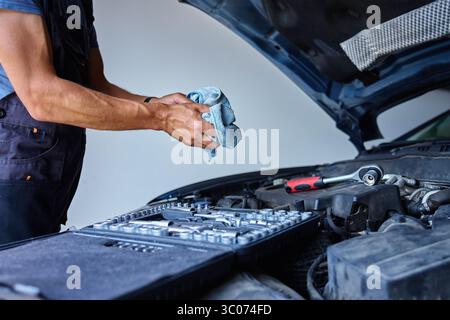 Auto mechanic cleaning dirty hands with cloth after completing car engine repair, standing near open car hood. Man finishing work in auto repair shop. Stock Photo