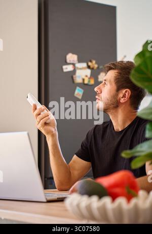Man working on laptop at home and using remote control to adjust temperature on air conditioner. Person struggling with heat while working at home off Stock Photo